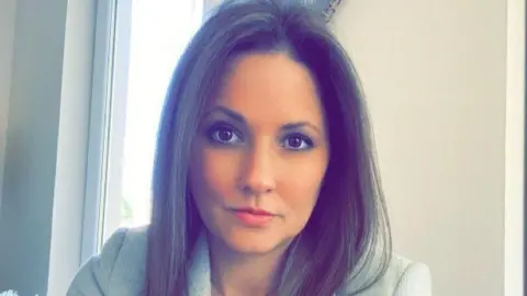 Annie Hirdman A woman with long brown hair stares directly at the camera. She has brown eyes and a serious expression on her face. The woman appears to have taken the selfie-style picture in a domestic setting, with white walls and a window visible in the background.