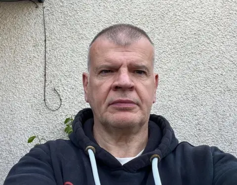 John Meiklejohn, a middle aged man dressed in a dark coloured hoodie, looks at the camera with a rendered wall of his home in the background.