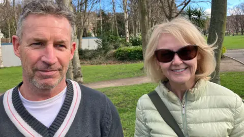 Les and Michelle in a park. There is grass and trees behind them. Les wears a grey jumper and white t-shirt. He has grey hair. Michelle wears a light green coat. She has blonde hair and is wearing sunglasses.