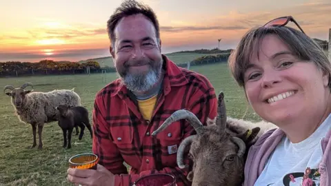 Ruth Meade Ruth Meade and husband Stuart in a field with a Loaghtan sheep in between them and another standing behind with a lamb. The sun is setting behind them.