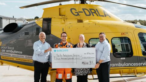 The Grace Trust presenting the cheque to The Air Ambulance Service, standing in front of a yellow helicopter