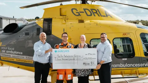 Wade & Tash McCroft The Grace Trust presenting the cheque to The Air Ambulance Service, standing in front of a yellow helicopter