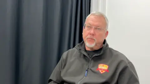 A man wearing a black top, with a grey goatie beard and glasses, with short white hair