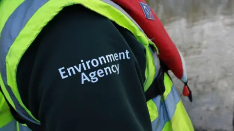 A close up of an Environment Agency employee's outfit. All that is visible is a high vis jacket and the shoulder of a black top with Environment Agency written on it in white.