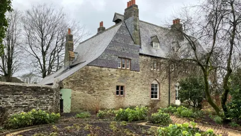 A substantial stone building with with a traditional slate roof and patterned  hanging slate to a gable end. There are hellebores in the garden and a tree with no leaves. 