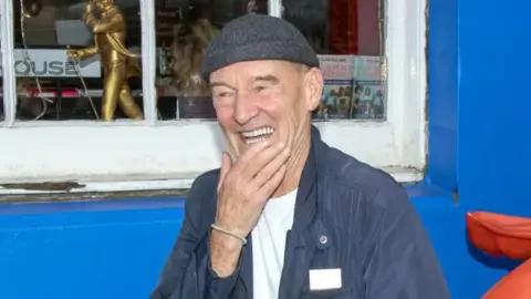 Getty Images David Hayman, in blue jacket, white T-shirt and grey wool hat, stands in front of a blue wall and white-framed window of a shop. He is laughing and his hand is resting on his chin.