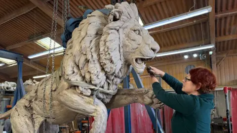 Tanya Russell A large lion that is being sculpted out of clay by a woman who is standing in front of it. The woman has short, red hair and is wearing a dark green top. She is holding a tool near the lion's mouth. The statue is in a large studio.