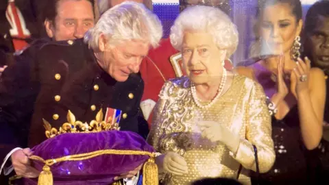 Martin Giles/BBC Bruno Peak standing beside Queen Elizabeth at the lighting of her Diamond Jubilee Beacon at the Mall, London. He is on the left and wearing a black jacket with gold buttons and holding a purple gold trimmed cushion with a crown on it. He is leaning towards the Queen, on the left and she is wearing a gold sequinned dress and speaking to him. There are people behind them.