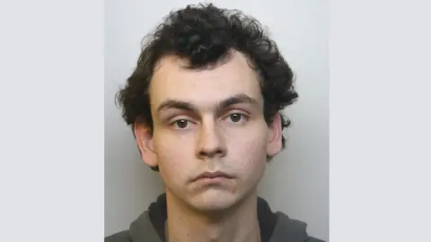 A police mugshot of Connor Chard, who is wearing a dark grey hoodie and standing against a plain grey background. He has black curly hair and bushy eyebrows, and is looking at the camera with a blank expression.