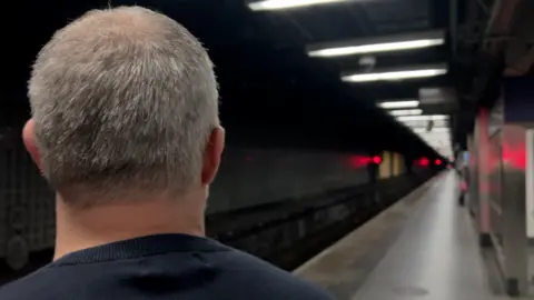 Grey-haired Keith Watson, seen from behind, on the platform of a railway station