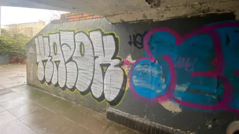 A large colourful piece of graffiti on a wall in a crumbling underpass. It is a wet day and the pavement has water on it.