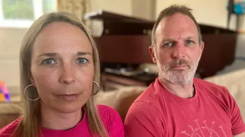 Maternity care campaigners Sarah and Jack Hawkins sitting next to each other on a sofa. They are both wearing pink t-shirts and looking directly at the camera.