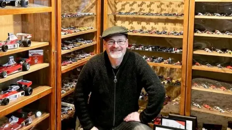 Sue Parton A man stands inside a room lined with wooden display cabinets filled with miniature racing cars. The shelves behind are densely arranged with detailed model vehicles in various colours and styles. The scene highlights a carefully curated collection showcased in a warm, museum‑like setting.