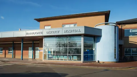 A photo of the front of Harrogate District Hospital. It is a brick building with silver-grey signage and a large window at the front, through which you can see some green chairs. 