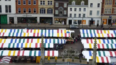 Aerial photo of Cambridge's Market Square. The stalls have canopys which are white and stripped colours and are in three long rows. Behind them are a row of shop.