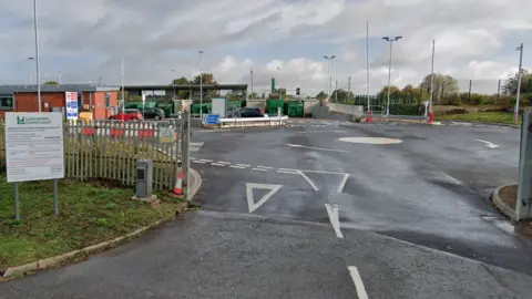 The entrance to a tip site from the road, with a roundabout just inside the site leading to waste bays