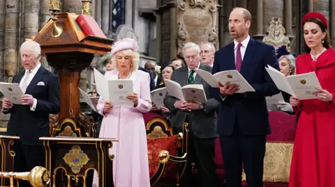 PA Media The King, Queen, Prince William and Catherine stand among a congregation of people inside a church as they sign from a hymn book.