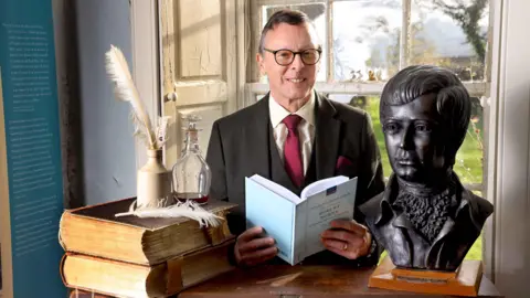 A man in glasses with a red tie and a brown jacket and cream shirt opens a book next to a bust of Robert Burns and some books and quill pens