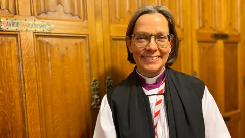 Bishop of Newcastle Helen Ann Hartley standing next to a wooden door. She wears her dog collar and a purple shirt, with a while and white robe. She had short dark bobbed hair and glasses.
