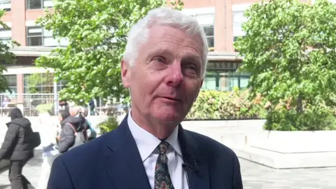 A man in a blue suit and patterned tie speaks in front of trees and white stone benches.