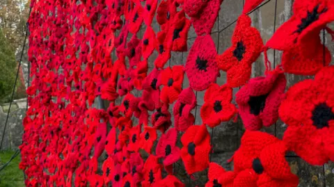 Liz Woodlands Hundreds of red knitted poppies that have been woven together attached to webbing on the side of the church building. 