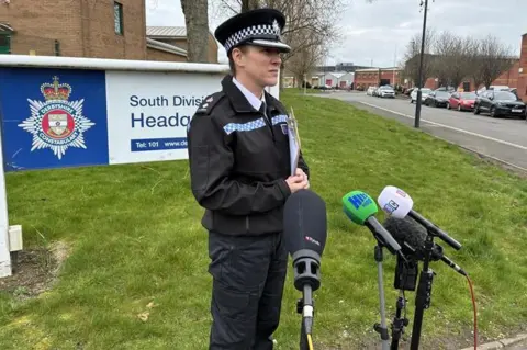 George Torr/BBC A woman in police uniform in infront of four microphones. 