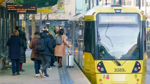 A yellow tram stops at a platform in Manchester and people on the platform prepare to get on