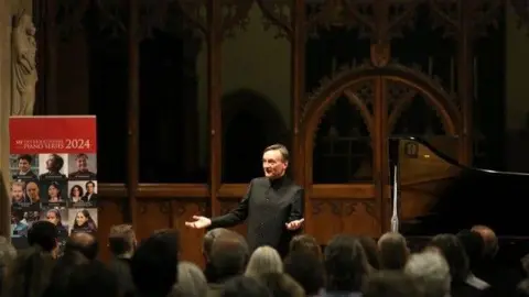 SJE Arts Sir Stephen Hough at SJE Arts event, He is talking to the audience. A banner of the Piano Series is seen at the side.