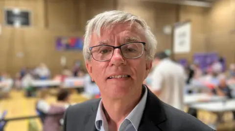 Steve Jarvis with short white hair and glasses, looking at the camera while wearing a dark grey jacket and a light grey shirt. There is an election count going on behind him in a sports hall.