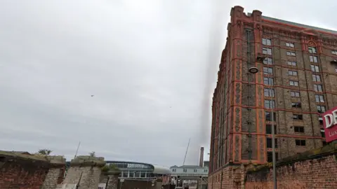 Google Image shows the redeveloped Tobacco warehouse on the Liverpool docks. It is a vast brick building more than 10 storeys high. To the left-hand side of the image, the Everton Hill Dickinson Stadium can be seen in the distance. 