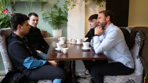 Sir Gareth Southgate sits at a wooden table in a cafe with three young men. They all have mugs in front of them