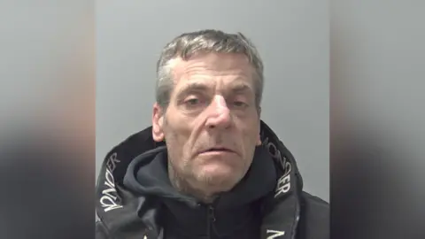 A man in a black hooded sweatshirt and black jacket. The collar of the jack has some white lettering on the inside. 