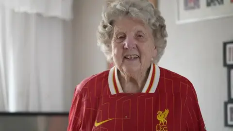 BBC Peggy Pickavant is smiling at the camera. She is wearing a Liverpool FC shirt and is standing in her living room in front of a TV with photographs hanging on the walls.