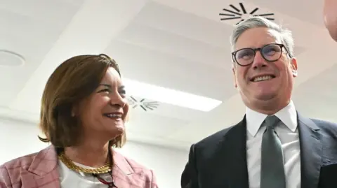 Prime Minister Sir Keir Starmer stands on the right of Wales' First Minister Eluned Morgan. He is wearing a dark suit and grey tie. She is wearing a pink checked jacket and necklace 