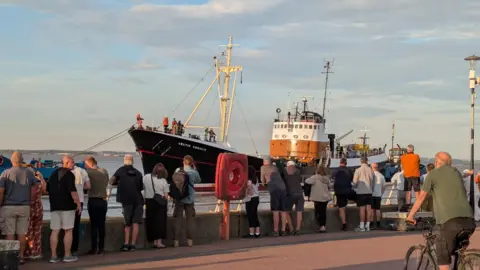 A long line of people are gathered along a wall as they look out at Arctic Corsair, a navy and white trawler that is making its way through the water.