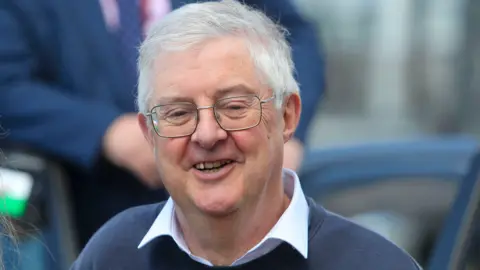 Mark Drakeford wearing a blue jumper, a shirt and glasses, smiling into a camera.