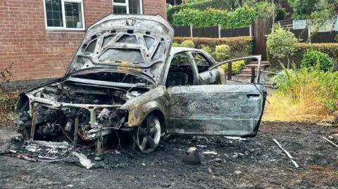 A burnt out shell of a car which was on fire. Debris and charred materials can be seen surrounding the vehicle.