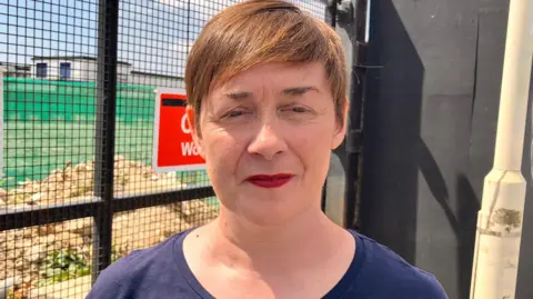 Sarah Williams is seen in a navy blue top stands outside a fenced construction site, with warning signs reading "Construction site" and "Keep out" visible behind her.