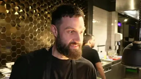 Alex Claridge, a man with a brown beard is wearing a black T-shirt and looking to the right of the frame while talking. He is standing in a restaurant kitchen.