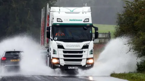 A lorry and car drive through flood water on a country road