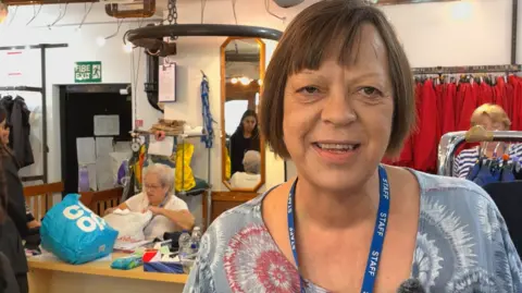 Marc Gaier/BBC A middle-aged woman with brown hair cut into a bob, wearing a grey and white patterned blouse and a blue lanyard, smiles at the camera. Behind her are clothes rails while volunteers are sorting clothes and serving customers. 