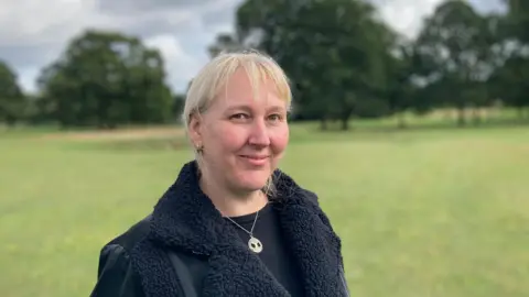 Laura Coffey/BBC Sammie Wakefield with short straight blond hair, smiling at the camera and wearing a black coat and silver necklace. She is standing in a park with trees in the background.