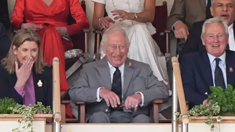 King Charles sitting in the stand at the horse show. There is a man and woman sitting either side. All of them are laughing at something which is out of shot.