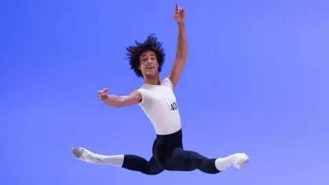 Gregory Batardon & Rodrigo Buas Jakob in full dance position, in the air, with his arms up and legs outstretched. He is wearing a white top with a number on it, black leggings, white socks and white dance shoes. He has short curly hair. 