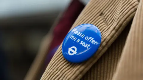 Transport for London A close-up of a blue "Please offer me a seat" badge pinned to the lapel of a tan corduroy jacket.