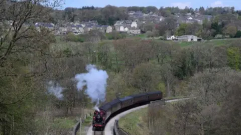 A steam train travels through a picturesque rural area, with trees, hills and houses all visible around and behind it.