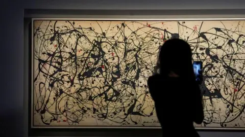 Reuters A person with long hair in silhouette is seen facing the artwork holding their mobile phone to video the painting. The artwork is long horizontally and the grey canvass appears a light tan colour under the gallery lights. It features black paint flicks and splashes all over it, together with some fewer red markings.