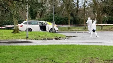 A white car with the driver's door open. The area is cordoned off by the police and there are forensic civil servants who run around the car. 