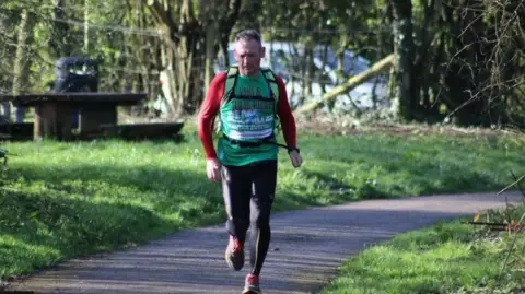 Gary McKee  Gary McKee running in a park with large green verges on either side and trees behind with a car in the distance.