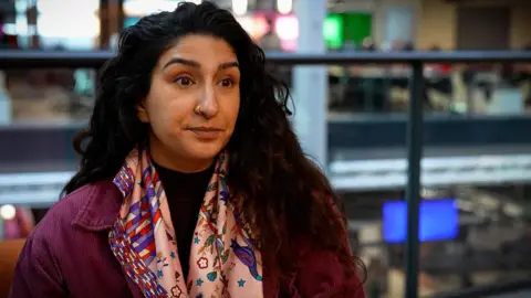 Trishna is sitting on an sofa in an office. She is looking across the camera, and has black hair and brown eyes. She is wearing a purple jacket and a patterned pink scarf